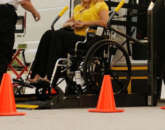 A person in a wheelchair uses a lift to board a van, assisted by a driver. Outdoor setting with cones.